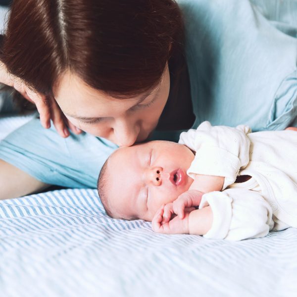 Mother kissing her newborn baby. Young beautiful mom lying at bed with a cute little sleeping child. New born baby's first days of life in family at home. Loving mother looks at asleep infant.